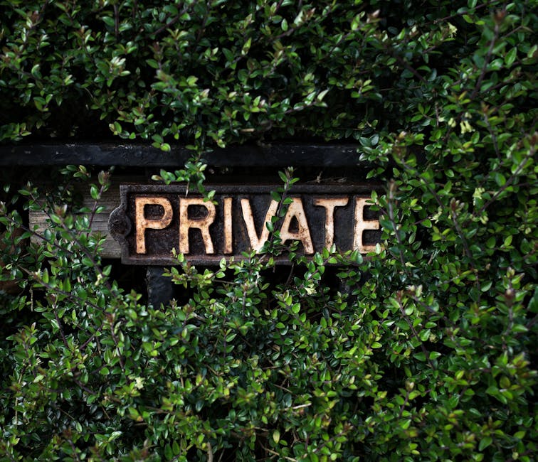 A close-up image of a weathered, rusty 'PRIVATE' sign mounted on a dark wooden board, partially obscured by dense, glossy green foliage of small, oval-shaped leaves. The lush shrubbery surrounds the s